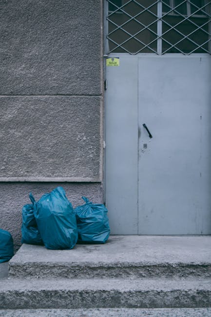 The image displays three blue rubbish bags placed on a concrete step outside a building, positioned to the left of a metal door with a black handle and a small lock. The bags appear to be made from a durable, slightly crinkled plastic material, and their bright blue color contrasts with the surrounding grey concrete and textured wall. The building's exterior wall is divided into two sections: the lower part is smooth with a light grey finish, while the upper section features a rough, darker grey finish. To the right of the rubbish bags, the door has a metal grille panel at the top, with a diamond pattern, and is secured with a black handle and locking mechanism. The scene is illuminated by natural daylight, with soft, even lighting highlighting the textures of the concrete, metal, and plastic. This setting suggests an environment suitable for private rubbish collection or independent waste handling, aligning with the context of affordable rubbish removal prices and services provided by Rubbish Removal Croydon.