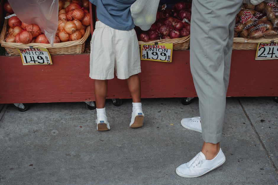 A close-up view of two individuals standing at a fruit and vegetable stall on a sidewalk. The child on the left wears a blue and yellow shirt with beige shorts, white socks, and brown shoes, facing the produce with their back partially visible. The adult on the right is dressed in light gray pants and white sneakers, also facing the display. The stall is constructed with a red-painted wooden base fitted with black wheels, and it displays baskets filled with onions, which are yellow and red in color, and partially covered with a transparent plastic sheet. Price tags showing the cost per kilogram are visible, with the onions priced at £1.49 and £1.99, respectively. The surrounding environment includes a concrete pavement, and the setup appears to be part of an outdoor market or grocery stand, which is relevant to independent food sourcing and waste management. The scene is lit naturally, with no additional lighting effects, emphasizing a neutral, everyday setting suitable for describing private and commercial produce displays related to waste and rubbish handling services.