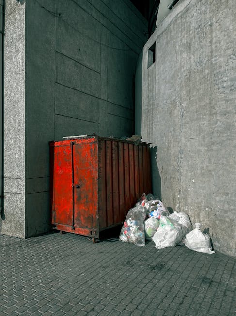 The image displays three blue rubbish bags placed on a concrete step outside a building, positioned to the left of a metal door with a black handle and a small lock. The bags appear to be made from a durable, slightly crinkled plastic material, and their bright blue color contrasts with the surrounding grey concrete and textured wall. The building's exterior wall is divided into two sections: the lower part is smooth with a light grey finish, while the upper section features a rough, darker grey finish. To the right of the rubbish bags, the door has a metal grille panel at the top, with a diamond pattern, and is secured with a black handle and locking mechanism. The scene is illuminated by natural daylight, with soft, even lighting highlighting the textures of the concrete, metal, and plastic. This setting suggests an environment suitable for private rubbish collection or independent waste handling, aligning with the context of affordable rubbish removal prices and services provided by Rubbish Removal Croydon.