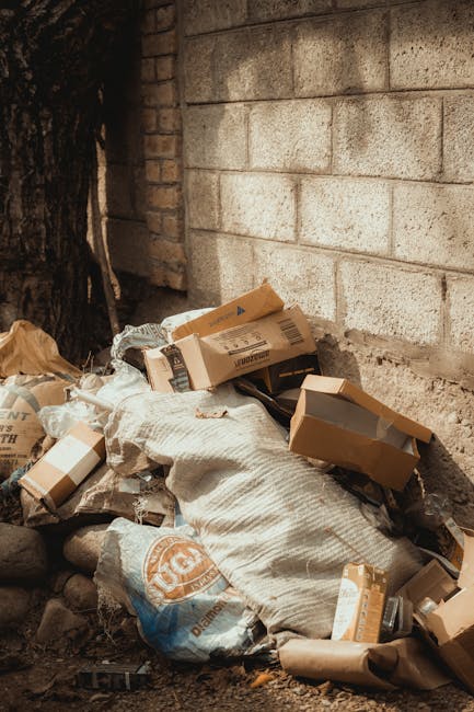 A pile of discarded packaging materials and rubbish, including flattened cardboard boxes, brown paper bags, and plastic bags, is situated against a brick wall and rough stone ground. The cardboard boxes vary in size, some partially opened or torn, with visible printed labels and barcodes. Behind the rubbish, a partially visible brick wall and a large tree trunk provide the backdrop. The scene appears to be an outdoor area, possibly a back alley or corner of a property, where waste has been accumulated for collection or disposal, highlighting an environment suitable for private rubbish removal services like those offered by Rubbish Removal Croydon. The overall setting is illuminated with natural light, casting minimal shadows on the mixed materials, emphasizing the textures of the cardboard, paper, and coarse ground beneath. The image underscores the need for proper waste management and alternative collection methods outside of local authority services.