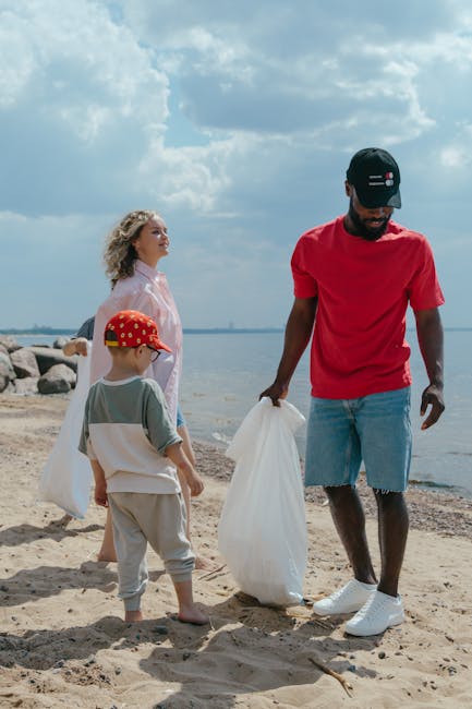 A group of three people, including a man and two children, are on a sandy beach near a body of water. The man, positioned on the right, is wearing a red T-shirt, light blue denim shorts, white sneakers, and a black cap with logo details, and he is holding a large white rubbish bag. The boy, in front of him, is wearing a beige and green T-shirt, light beige pants, and a red cap with yellow details, and is holding another white bag. The woman, standing to the left, has curly blonde hair and is dressed in a light pink outfit, gazing out towards the water with a calm expression. Behind them are large rocks forming a shoreline, with a cloudy sky overhead indicating an overcast day. The scene suggests a beach cleanup activity, aligning with the context of rubbish removal services, emphasizing private collection and environmental care in outdoor settings. Rubbish Removal Croydon occasionally supports such on-site clearance activities through alternative waste handling methods.