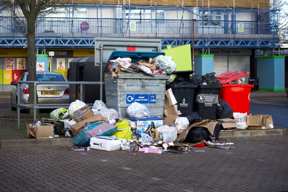 A pile of discarded packaging materials and rubbish, including flattened cardboard boxes, brown paper bags, and plastic bags, is situated against a brick wall and rough stone ground. The cardboard boxes vary in size, some partially opened or torn, with visible printed labels and barcodes. Behind the rubbish, a partially visible brick wall and a large tree trunk provide the backdrop. The scene appears to be an outdoor area, possibly a back alley or corner of a property, where waste has been accumulated for collection or disposal, highlighting an environment suitable for private rubbish removal services like those offered by Rubbish Removal Croydon. The overall setting is illuminated with natural light, casting minimal shadows on the mixed materials, emphasizing the textures of the cardboard, paper, and coarse ground beneath. The image underscores the need for proper waste management and alternative collection methods outside of local authority services.