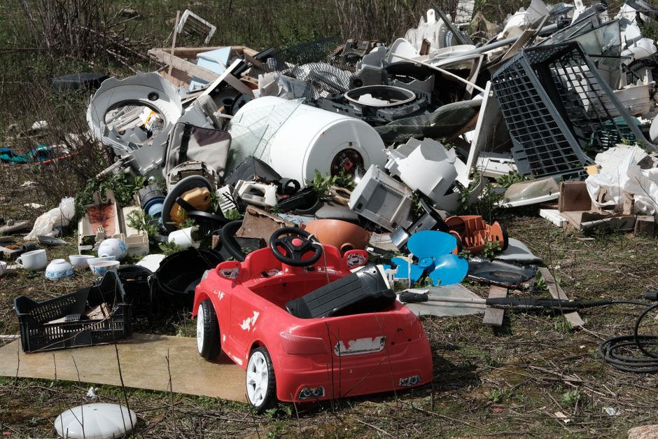 The image displays a large pile of assorted household waste and discarded items situated outdoors on a patch of uneven ground with some grass and sparse vegetation. Prominent in the foreground is a small red plastic children's ride-on car with minor scuffs and peeling paint on its exterior, positioned near the bottom left corner. Surrounding the car, various broken household objects are scattered, including plastic containers, bowls, and a black plastic crate. In the middle and background, there is a chaotic mound of larger appliances and rubbish components, such as a white washing machine drum, ceiling or exhaust fans, plastic storage baskets, and electronic parts, all partially covered with dirt, leaves, and small twigs. The scene suggests an informal dumping site or bulk waste clearance area, with no visible signage, fences, or containment measures, aligning with private waste handling or alternative rubbish removal services sometimes employed for large or bulky household debris, as might be managed by companies like Rubbish Removal Croydon.