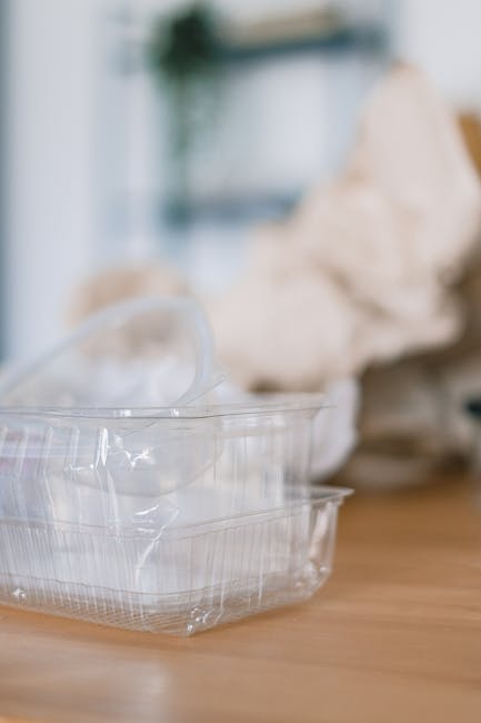 A clear plastic food container with a transparent lid and a second, slightly larger container underneath, placed on a wooden surface. The containers are made of rigid, glossy plastic with a ridged design on the sides. In the background, there is a blurred view of crumpled paper or tissue and a faint hint of a plant and shelving, suggesting an indoor setting such as a kitchen or dining area. The lighting is soft and natural, highlighting the transparency and texture of the plastic containers. The image portrays a scene related to waste disposal or recycling practices, aligning with independent rubbish removal or on-site clearance services offered by Rubbish Removal Croydon.