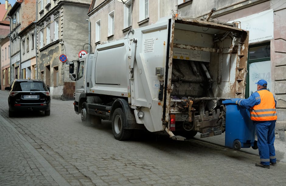 A waste collection worker dressed in a blue uniform with an orange reflective vest is positioned on a narrow cobblestone street, actively emptying a blue waste bin into the rear hopper of a large white rubbish collection truck. The truck, parked alongside an older row of multi-story buildings with a mix of stone and plaster facades, shows signs of rust and wear on its metal parts, particularly around the open hopper area. The scene is illuminated by natural daylight, highlighting the textured surfaces of the cobbled pavement and the weathered exterior walls. Behind the worker, a black vehicle is parked parallel to the truck, and there are traffic restriction signs mounted on poles attached to the buildings. This setting depicts an instance of private or independent waste collection, characteristic of scheduled rubbish removal services provided by companies like Rubbish Removal Croydon, aligned with the context of same-day or on-site clearance for urban waste management in Croydon town centre.