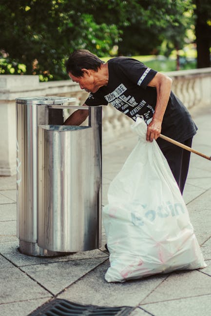 A middle-aged woman with short dark hair, dressed in a black t-shirt with white text and dark trousers, is bent over, inserting rubbish into a cylindrical metal recycling bin located on a paved sidewalk. She holds a large, white plastic bag filled with waste in her left hand and uses her right hand to lift a piece of rubbish into the open top of the bin. The bin features a smooth, reflective stainless steel finish with a rounded opening at the top. The scene is set outdoors in a green urban environment, with a low stone wall and lush foliage visible in the background, suggesting a park or town centre setting. The lighting is natural, indicating daytime, and the focus is on the woman actively participating in waste disposal, which aligns with private rubbish removal or on-site clearance services provided by Rubbish Removal Croydon.