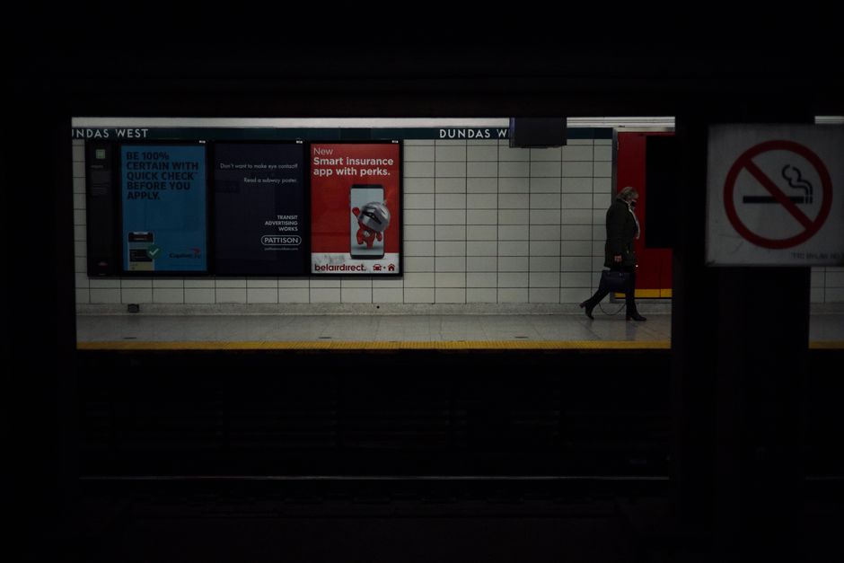 A person walking along the platform of Duda Dudas West underground station, which features white tiled walls with advertising posters, including one for smart insurance apps and another promoting transit services. The individual is dressed in dark clothing, with a face mask, and is carrying a black bag in one hand while walking towards a red door at the end of the platform. The platform is separated from the dark tunnel area in the foreground by a protective barrier, and a 'no smoking' sign is visible on the right side of the image. The scene is lit with artificial lighting, highlighting the clean, tiled surface and the advertisements, creating a contrast between the brightly illuminated platform and the darker tunnel opening, which could relate to the context of urban transit and the importance of maintaining cleanliness and order in public transport environments, reflecting a focus on efficient waste management and waste disposal services in busy transit hubs.