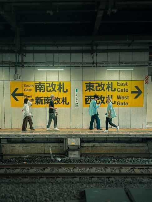 In an underground train station, five people are walking along the platform, all wearing casual clothing and face masks. The background features a white tiled wall with large yellow directional signs displaying black and red text in both English and Chinese. The signs indicate directions to the South Gate, East Gate, West Gate, and New Platform, with arrows pointing left and right. The platform surface appears to be concrete with tactile paving near the edge for safety. Overhead, the station's infrastructure includes exposed piping, lighting fixtures, and structural beams painted in neutral tones, creating a utilitarian environment typical of urban transit systems. The lighting is relatively bright, casting soft shadows on the platform, highlighting the movement of the commuters. The scene captures a moment of routine transit activity, where passengers are navigating their way to different station exits or transfer points, embodying the practical aspect of public transportation rather than private waste disposal or rubbish handling, subtly aligning with the theme of organized infrastructure maintenance.