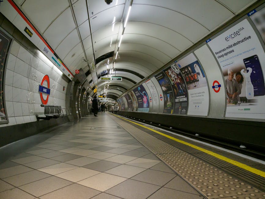 A person walking along the platform of Duda Dudas West underground station, which features white tiled walls with advertising posters, including one for smart insurance apps and another promoting transit services. The individual is dressed in dark clothing, with a face mask, and is carrying a black bag in one hand while walking towards a red door at the end of the platform. The platform is separated from the dark tunnel area in the foreground by a protective barrier, and a 'no smoking' sign is visible on the right side of the image. The scene is lit with artificial lighting, highlighting the clean, tiled surface and the advertisements, creating a contrast between the brightly illuminated platform and the darker tunnel opening, which could relate to the context of urban transit and the importance of maintaining cleanliness and order in public transport environments, reflecting a focus on efficient waste management and waste disposal services in busy transit hubs.